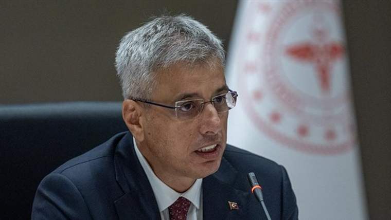 Mikrofon başında konuşan gözlüklü bir erkek, takım elbise ve kravat takıyor; arka planda kırmızı beyaz amblemli pano bulunuyor.