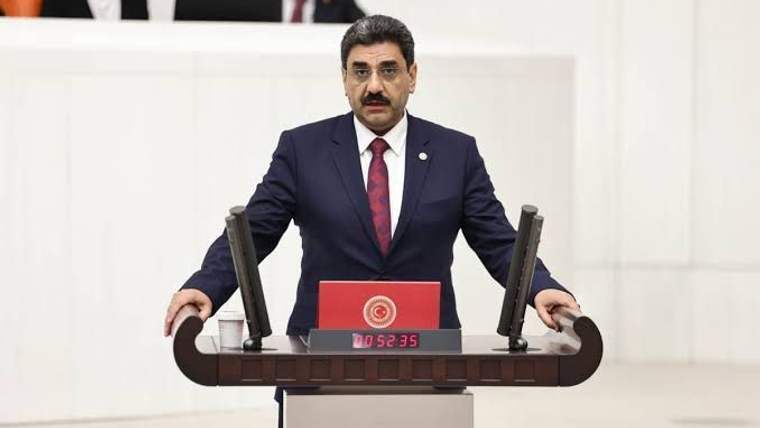 Kürsüde duran, koyu renk takım elbiseli ve kırmızı kravatlı bir erkek; önünde iki mikrofon bulunuyor.