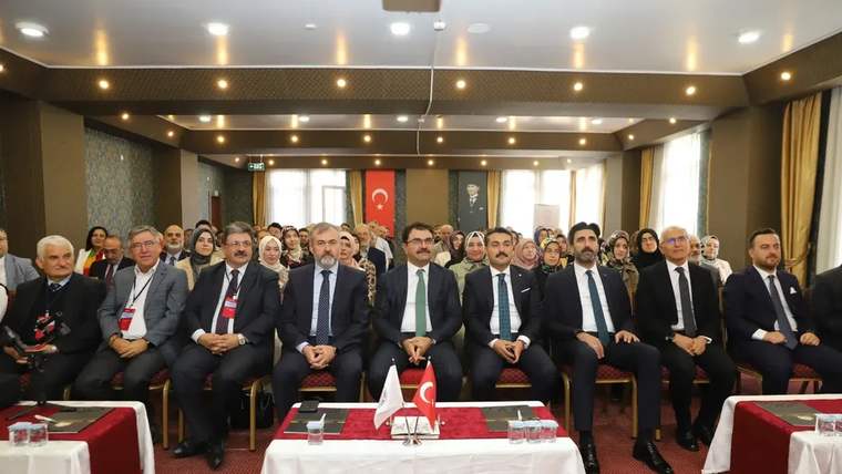 Toplantı salonunda takım elbiseli kişiler, Türk bayraklarının bulunduğu masaların önünde sıralı oturuyor.