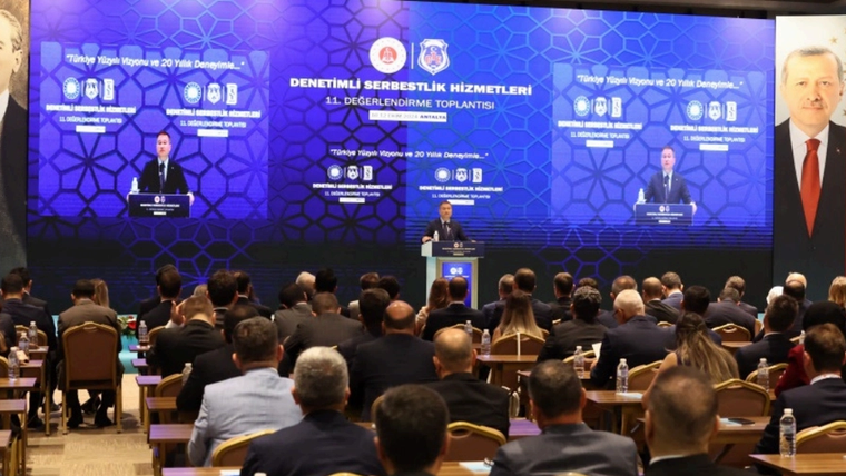 Bir toplantı salonunda, sahnede bir kişi konuşma yapıyor, dinleyiciler dikkatle izliyor.