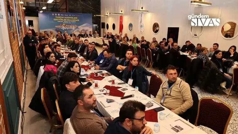 Toplantı salonunda uzun masalarda oturan kalabalık grup ve arka planda turizm temalı pankart görülüyor.