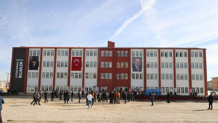 Kırmızı-beyaz cepheli okul binasının önünde insanlar toplanmış; girişte Türk bayrağı ve portreler asılı.