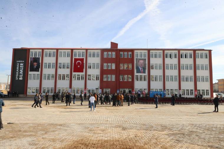 Kırmızı-beyaz cepheli okul binasının önünde insanlar toplanmış; girişte Türk bayrağı ve portreler asılı.