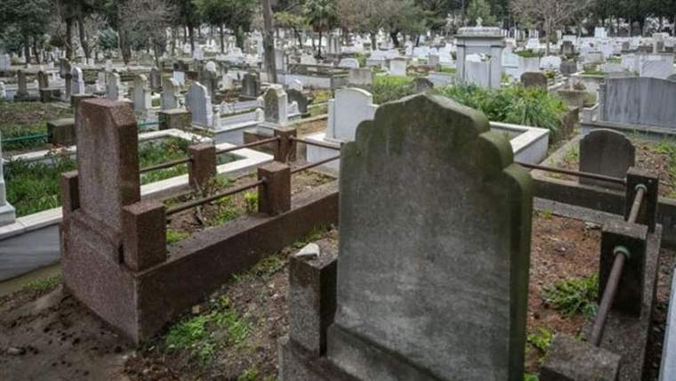 Bir mezarlık alanında çeşitli mezar taşları ve yeşil bitki örtüsü görünmektedir.