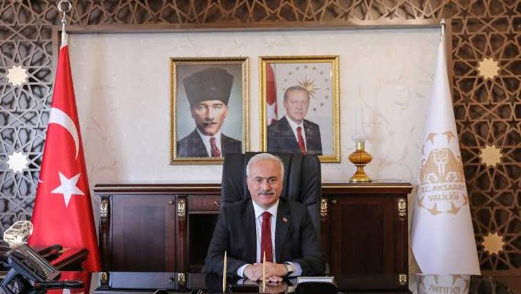 Bir masa arkasında oturan bir kişi, arkasında iki portre ve Türk bayrağı var.