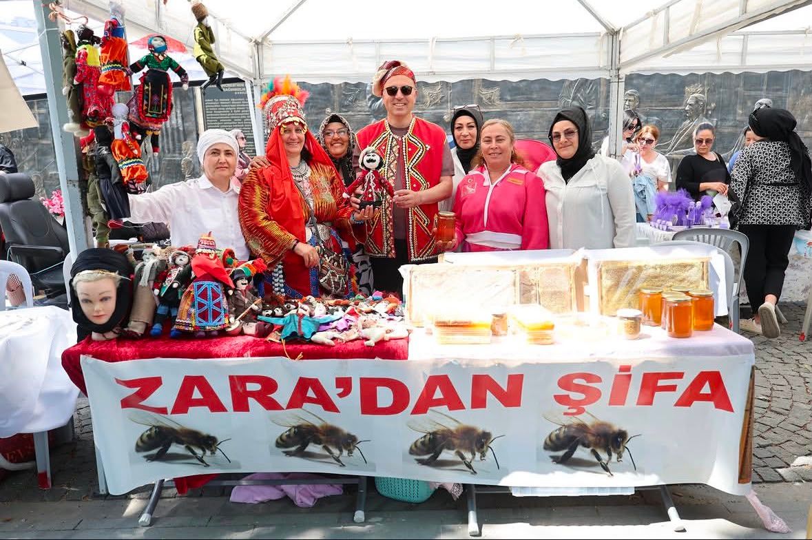 Çadırdaki stantta yöresel kıyafetli kişiler, el işi ürünler ve kavanozlar sergileniyor; masada “Zara’dan Şifa” yazıyor.