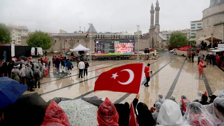 Yağmur altında, Türk bayrağı tutan kalabalık, dev ekranda Dünya Kupası maçını izliyor.