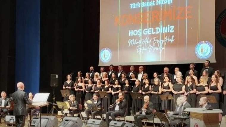Sahnede koro halinde duran şarkıcılar, önlerinde oturan saz sanatçıları ve onları yöneten şef görülüyor.