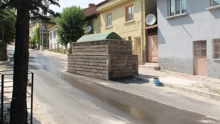 Yol ortasında duran taş yapıda bir mezar, çevresinde ağaçlar ve binalar var.