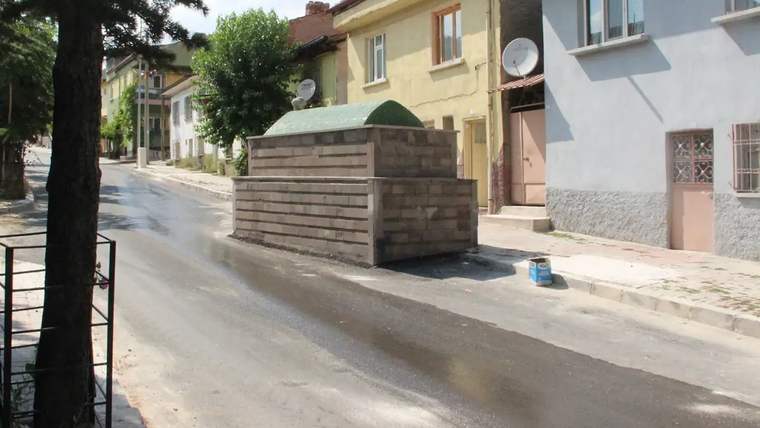 Yol ortasında duran taş yapıda bir mezar, çevresinde ağaçlar ve binalar var.