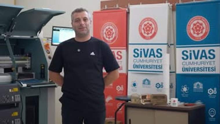 Bir kişi, baskı makineleri ve üzerinde Sivas Cumhuriyet Üniversitesi yazan tanıtım panolarının önünde duruyor.