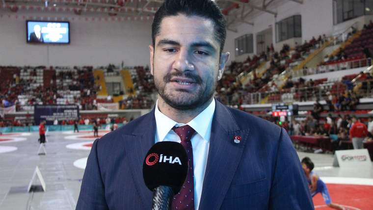 Takım elbiseli bir kişi, spor salonunda elindeki mikrofonla konuşuyor; arkasında minder ve tribünler görülüyor.