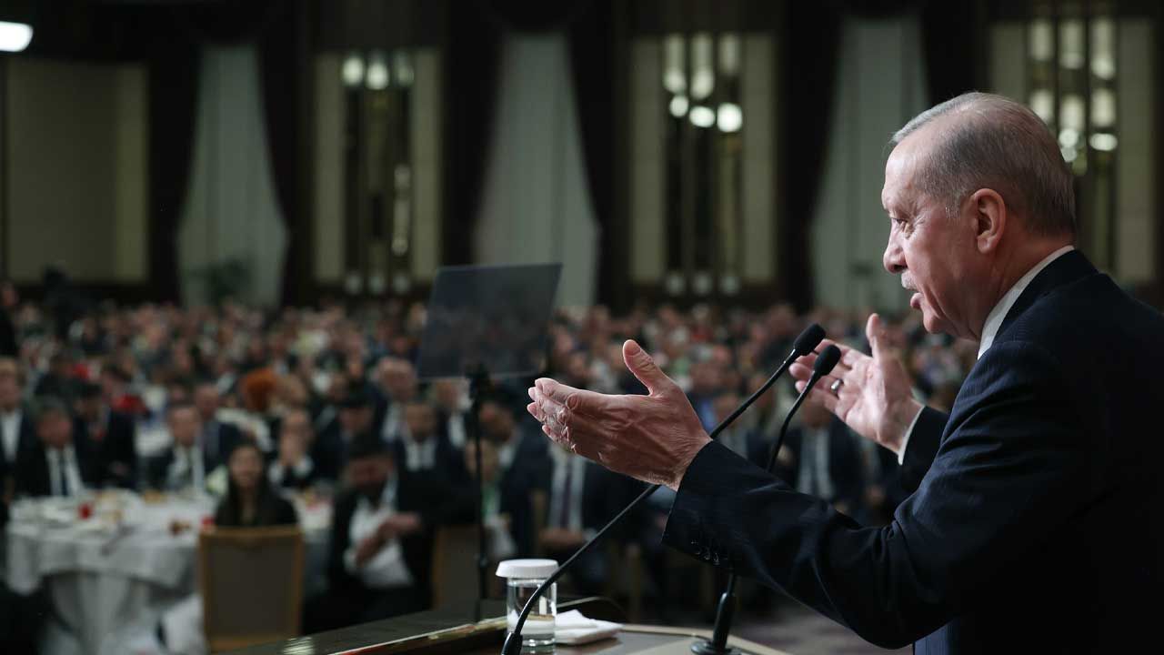 Mikrofon önünde konuşan bir kişi, kalabalık salona doğru ellerini uzatırken görülüyor.