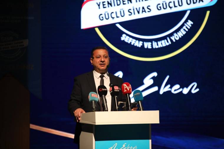 Kürsüde konuşan bir kişi, önündeki çok sayıda mikrofonla koyu renkli bir fonda duruyor.