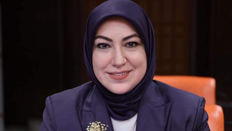 Başında başörtüsü olan bir kadın, gülümseyerek kameraya bakıyor. Arka planda koyu renkli bir ortam var.