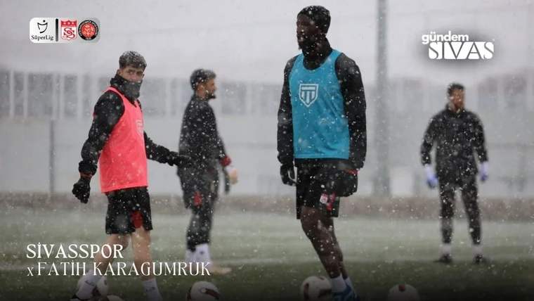 Yağış altında futbol sahasında duran, mavi ve kırmızı yelekli oyuncular ile arkadaki iki kişi görülüyor.