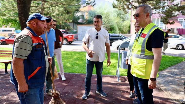 Bir adam elinde süpürge ile duruyor, çevresinde birkaç kişi var. Arka planda ağaçlar ve park alanı görünmekte.