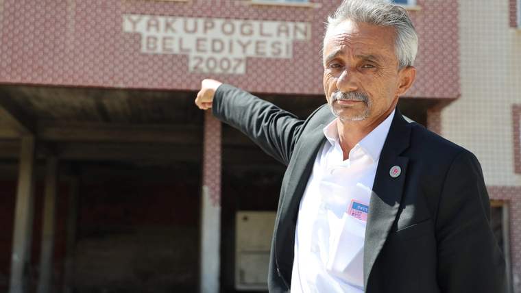 Bir kişi, arkasında 'Yakupoğlu Belediyesi 2007' yazılı bir bina ile işaret ediyor.