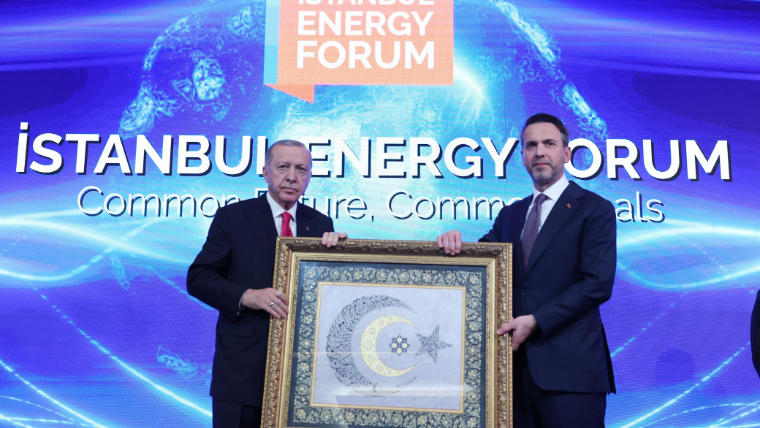 İki kişi, elinde çerçeveli bir tablo tutarak sahnede duruyor. Arkada İstanbul Enerji Forumu yazılı bir pankart var.