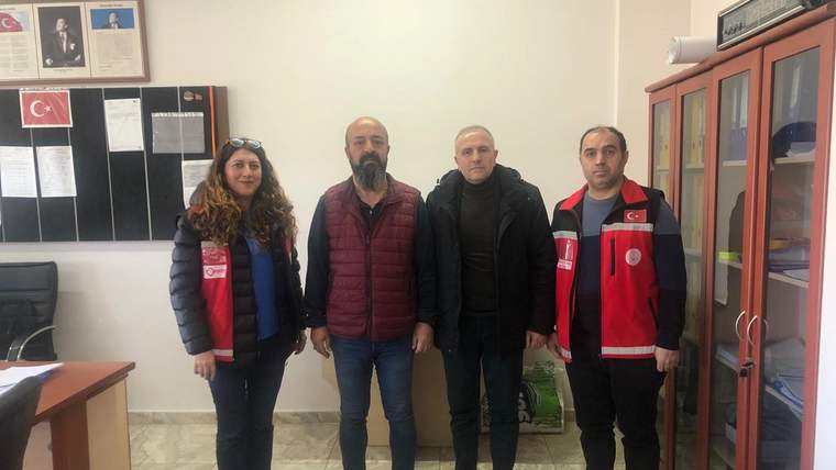 Ofis ortamında, ikisi kırmızı yelekli dört kişi panolar ve dolapların önünde yan yana duruyor.