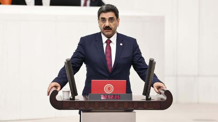 Kürsüde konuşan bir kişi, önünde mikrofonlar ve bir saat var.