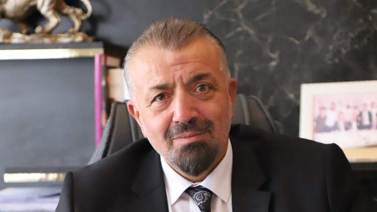 Takım elbiseli, kravatlı ve sakallı bir erkek, masa arkasında kameraya doğru oturuyor.