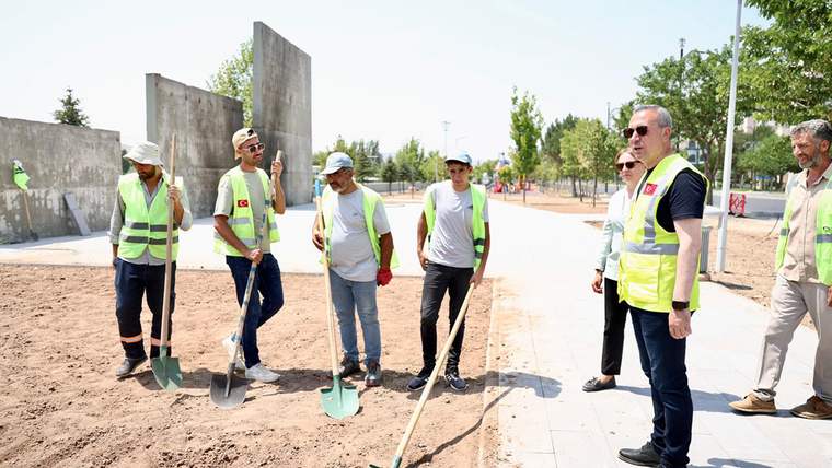 Park alanında kürek ve tırmık taşıyan işçilerle, fosforlu yelekli iki kişi yürüyüş yolunun yanında duruyor.