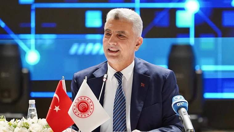 Masada mikrofon önünde konuşan bir kişi, Türk bayrakları ve çiçeklerle birlikte görülüyor.