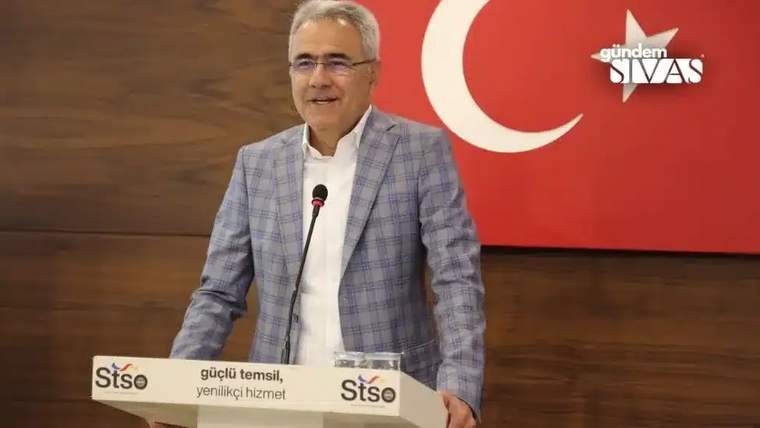 Bir kişi, arkasında Türk bayrağı bulunan bir kürsüde konuşuyor.