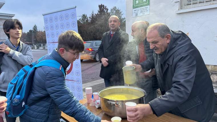 Bir adam, sıcak çorba dağıtırken, önünde bir öğrenci bekliyor; arka planda başka insanlar ve araçlar görünüyor.
