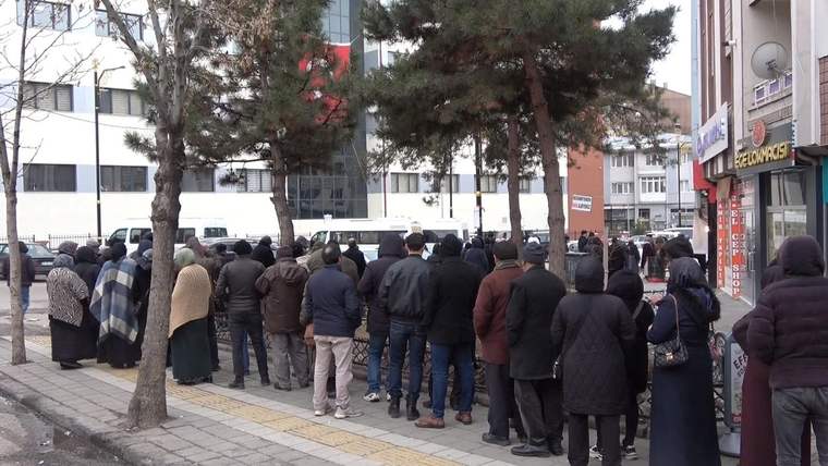 Uzun bir kuyrukta bekleyen kalabalık, ağaçların gölgesinde duruyor.