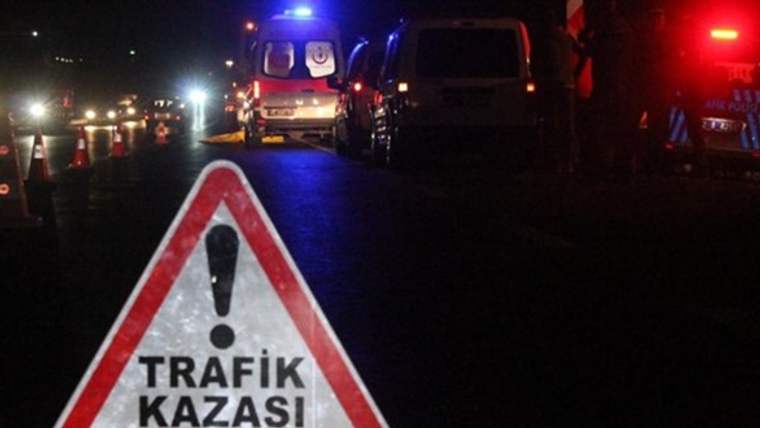 Gece karanlığında, üzerinde “TRAFİK KAZASI” yazan uyarı levhası, ışıkları yanan araçların önünde duruyor.