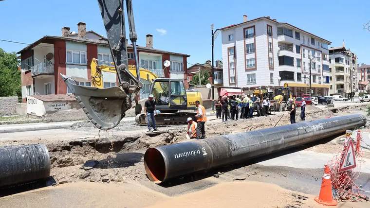 Şehir caddesinde kazı çalışması yapan iş makineleri, işçiler ve yola bırakılmış büyük su boruları görülüyor.