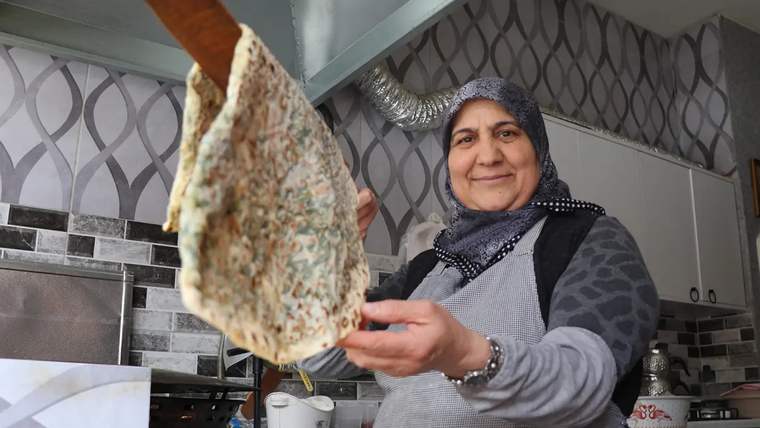 Başörtülü bir kadın, mutfakta spatulayla pişmiş gözlemeyi kameraya doğru tutuyor.