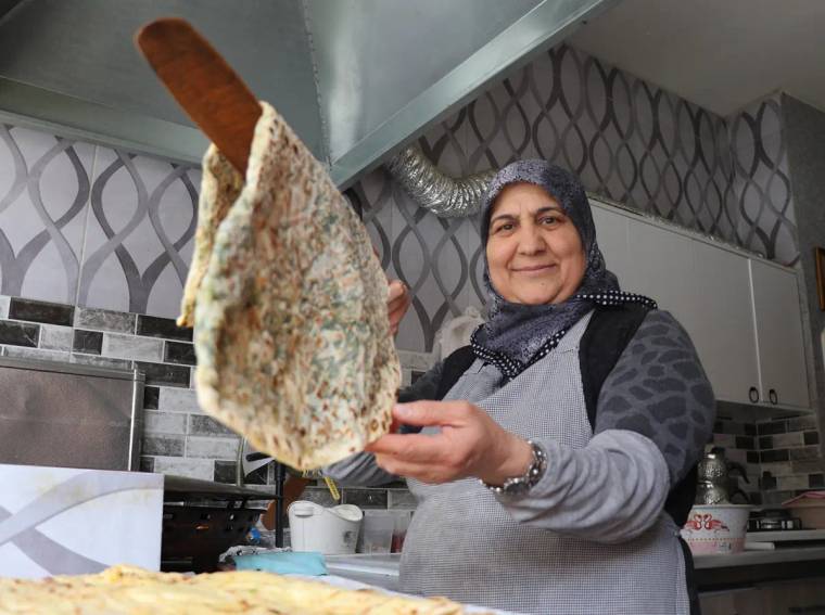 Başörtülü bir kadın, mutfakta spatulayla pişmiş gözlemeyi kameraya doğru tutuyor.