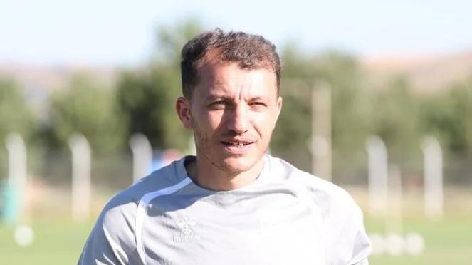 Antrenman alanında, açık havada gülümseyen bir futbolcu, gri antrenman kıyafeti giymekte.