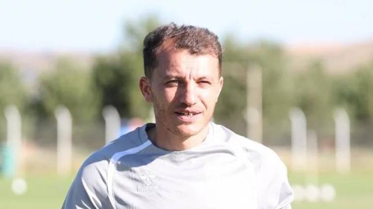 Antrenman alanında, açık havada gülümseyen bir futbolcu, gri antrenman kıyafeti giymekte.