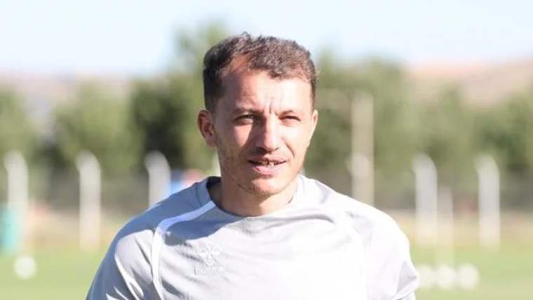 Antrenman alanında, açık havada gülümseyen bir futbolcu, gri antrenman kıyafeti giymekte.