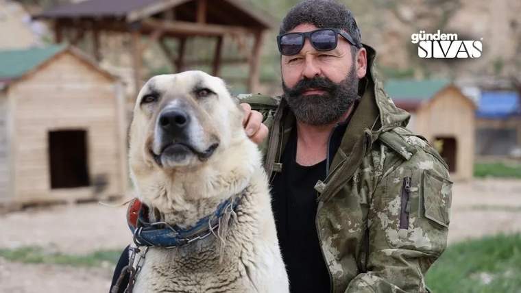 Kamuflaj desenli mont giyen sakallı bir kişi, büyük beyaz bir köpekle kulübelerin önünde duruyor.