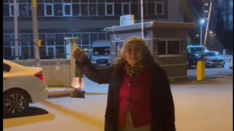 Bir kadın, elinde bir fener tutarak sokakta duruyor. Arkada binalar ve park halindeki araçlar görünmekte.