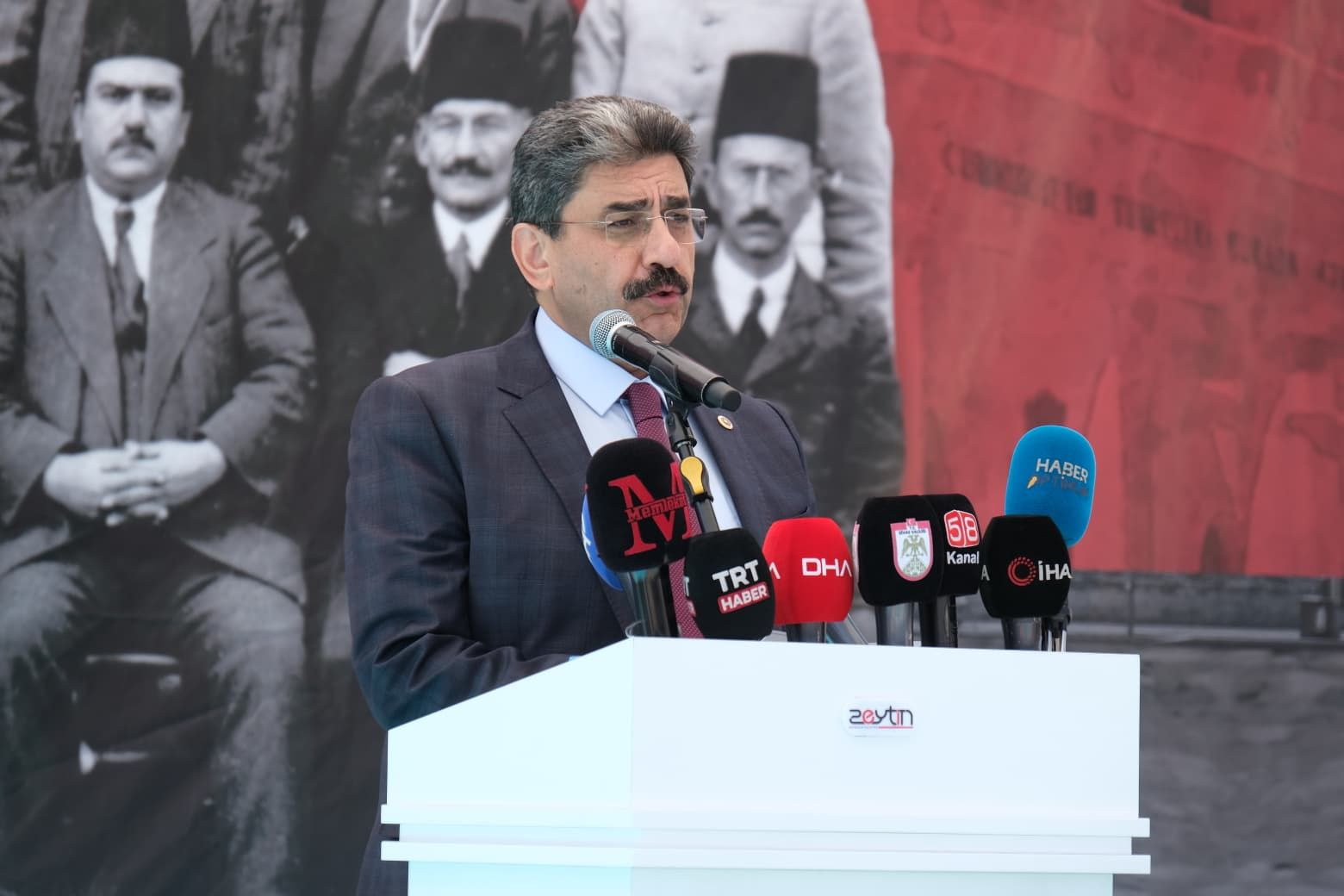 Takım elbiseli bir kişi, çok sayıda mikrofonun bulunduğu kürsüde konuşurken arkasında tarihi bir fotoğraf görülüyor.