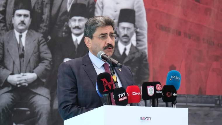 Takım elbiseli bir kişi, çok sayıda mikrofonun bulunduğu kürsüde konuşurken arkasında tarihi bir fotoğraf görülüyor.