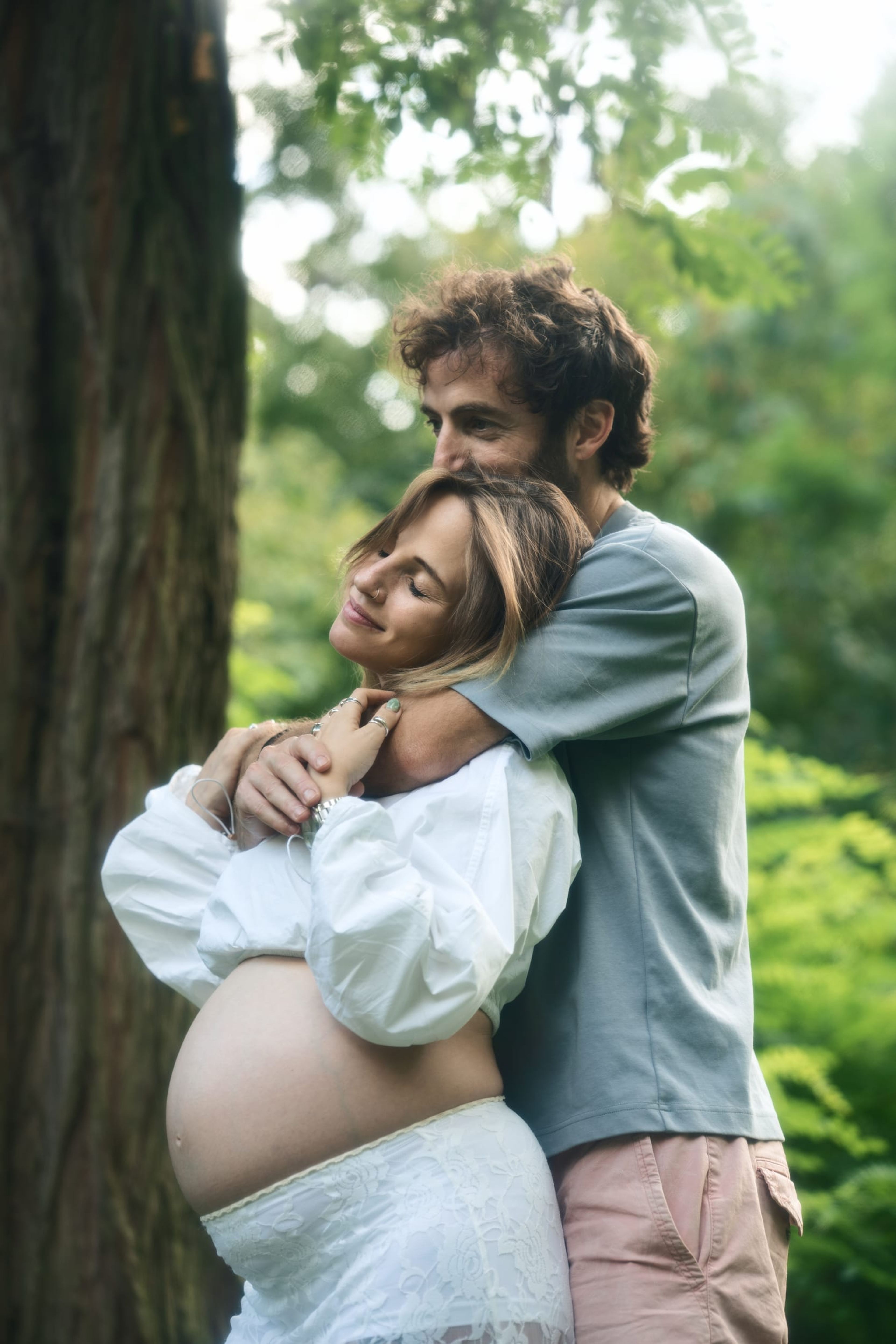 Partner holding the expectant mother against a tree, both with their eyes closed