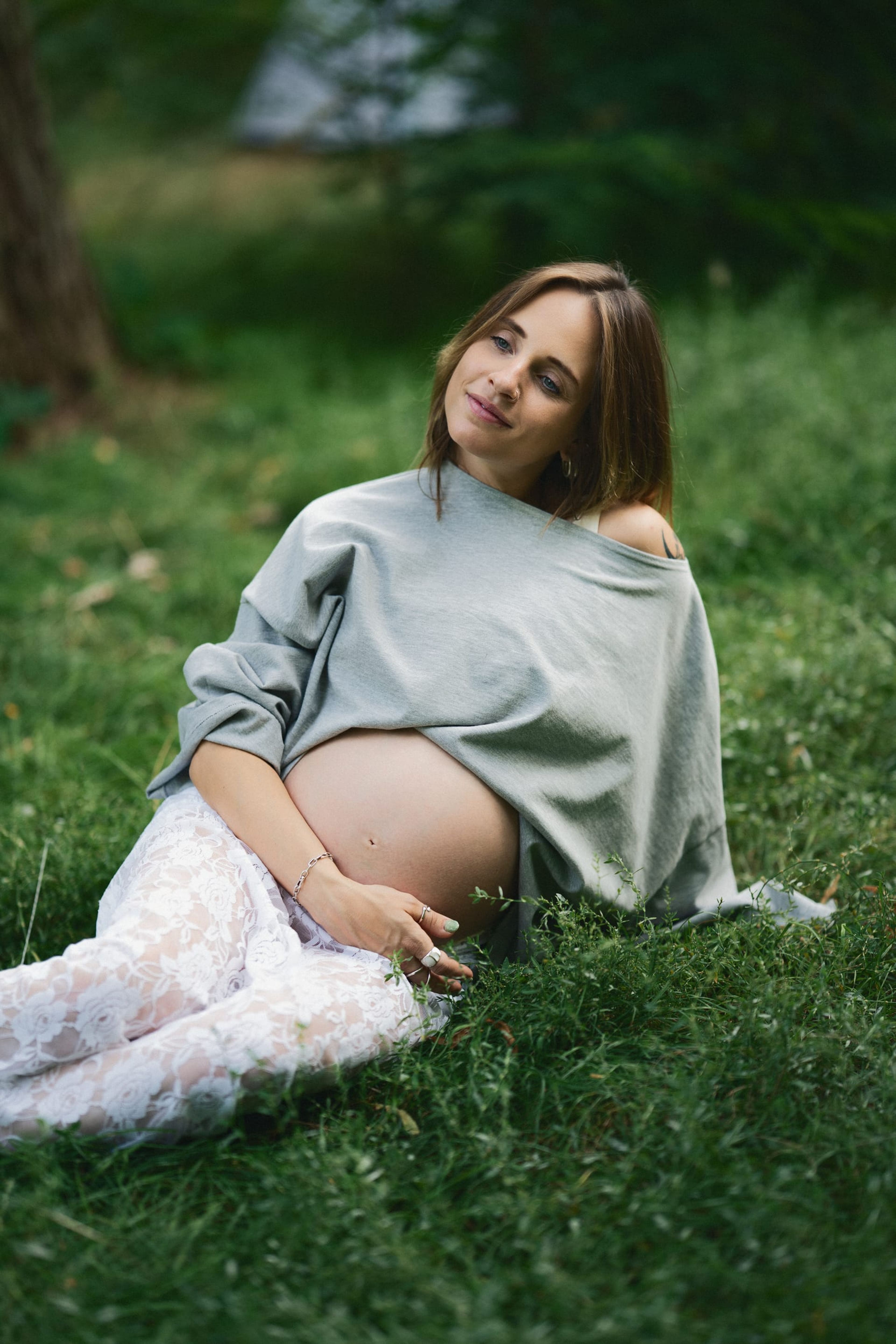 Expectant mother sitting in the grass in an off-shoulder sweater, looking to camera