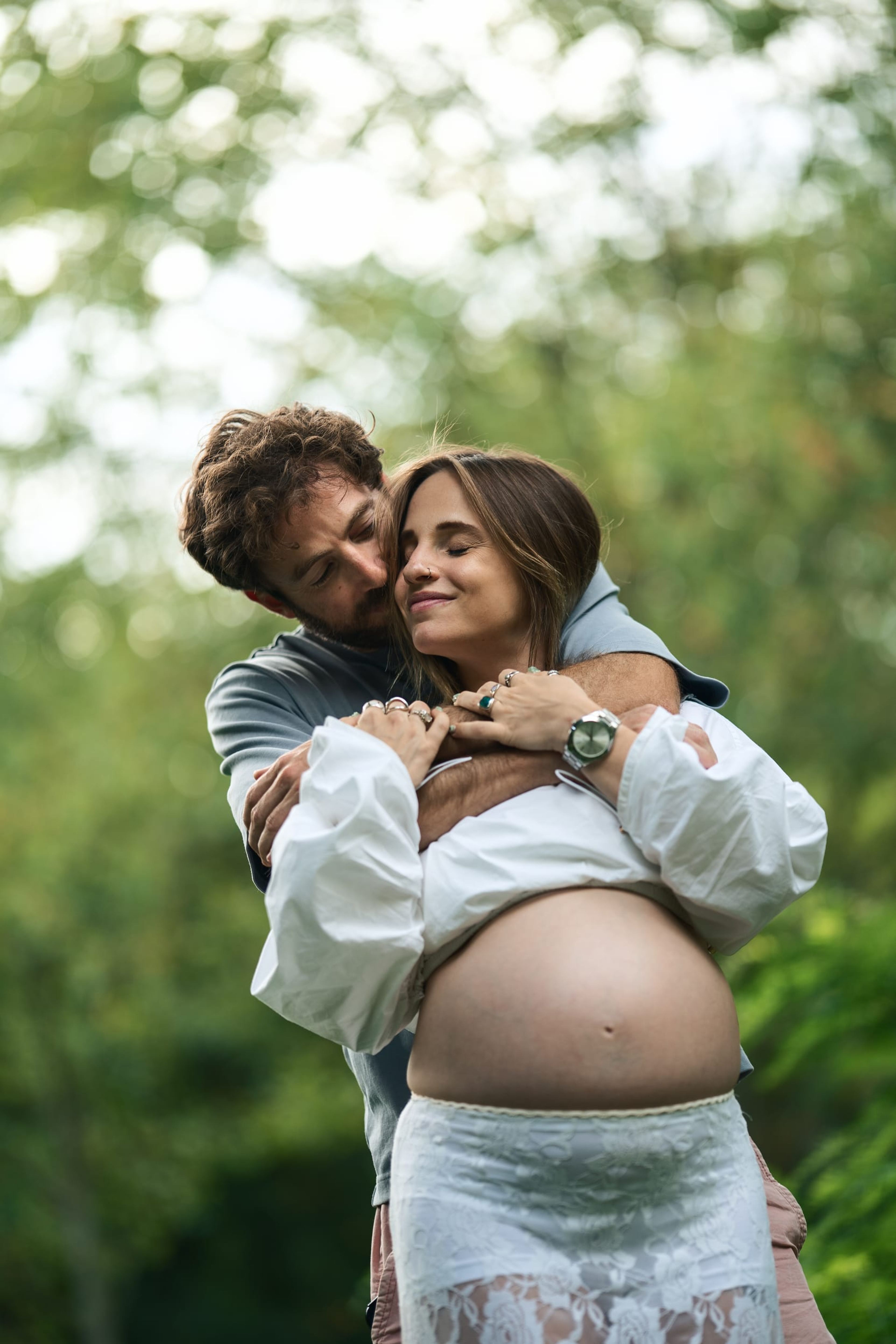 Partner embracing the expectant mother from behind in dappled light