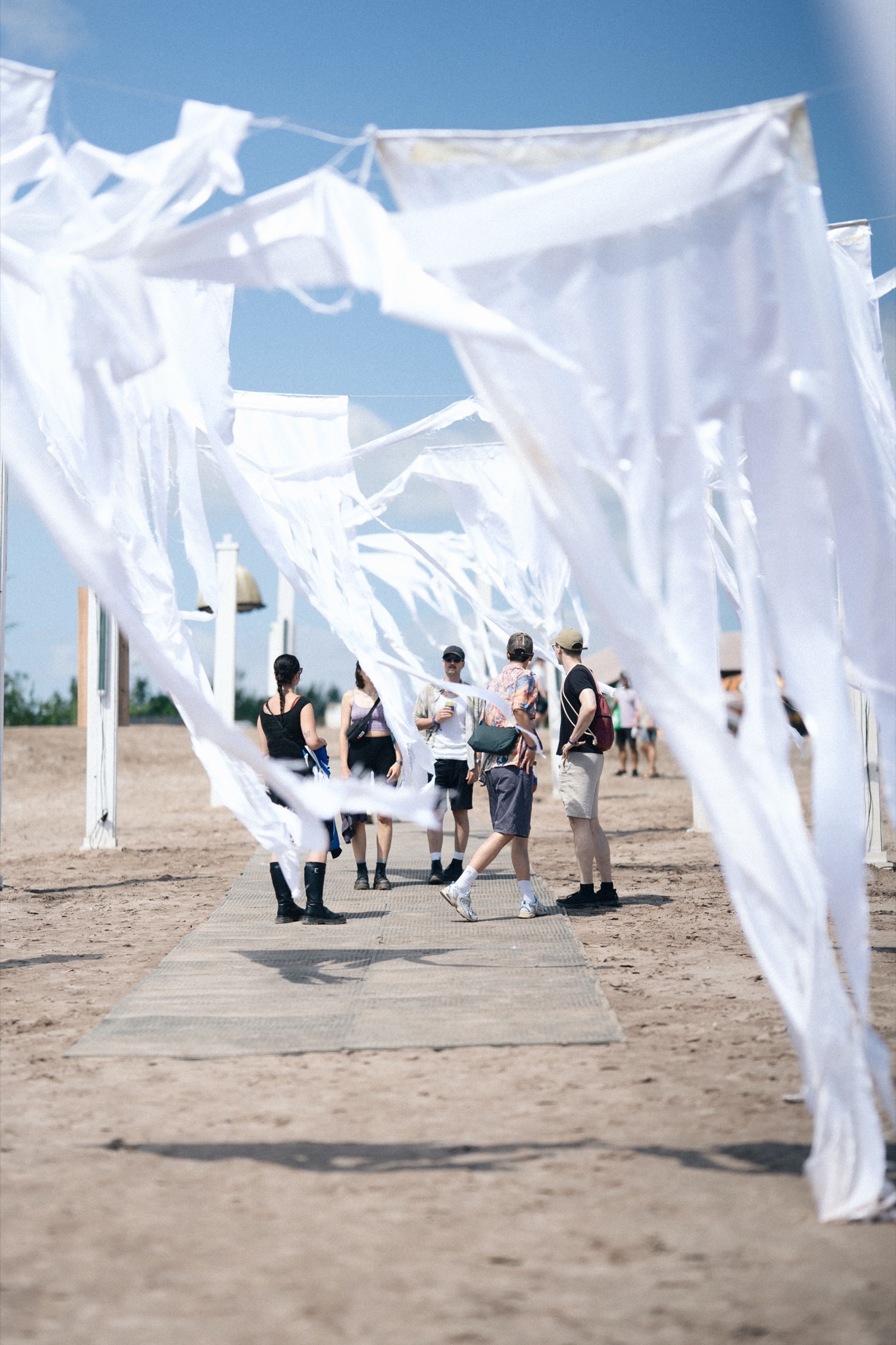 White fabric banners billowing over a sandy walkway between festival-goers