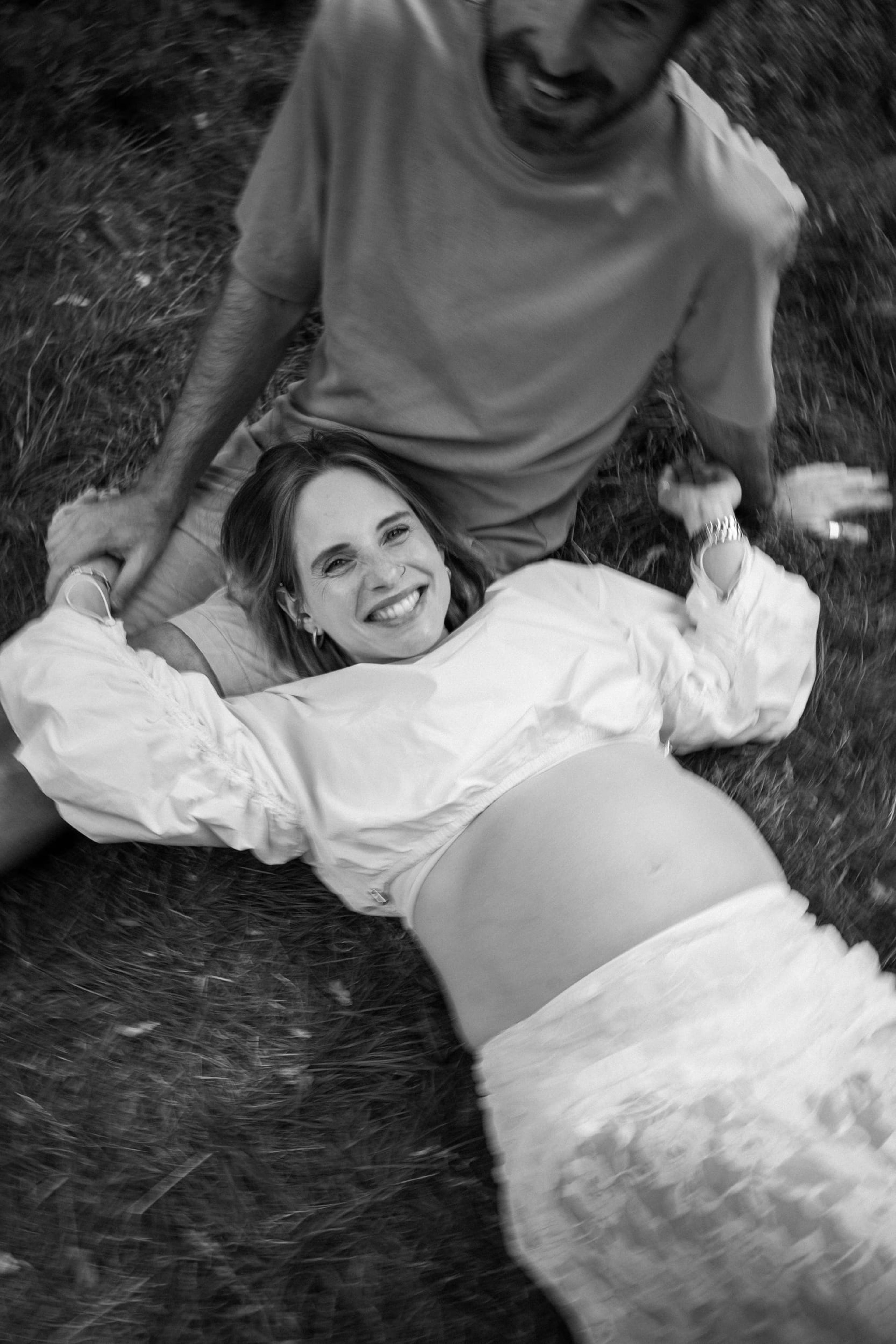 Black-and-white overhead frame of the expectant couple lying in the grass, laughing
