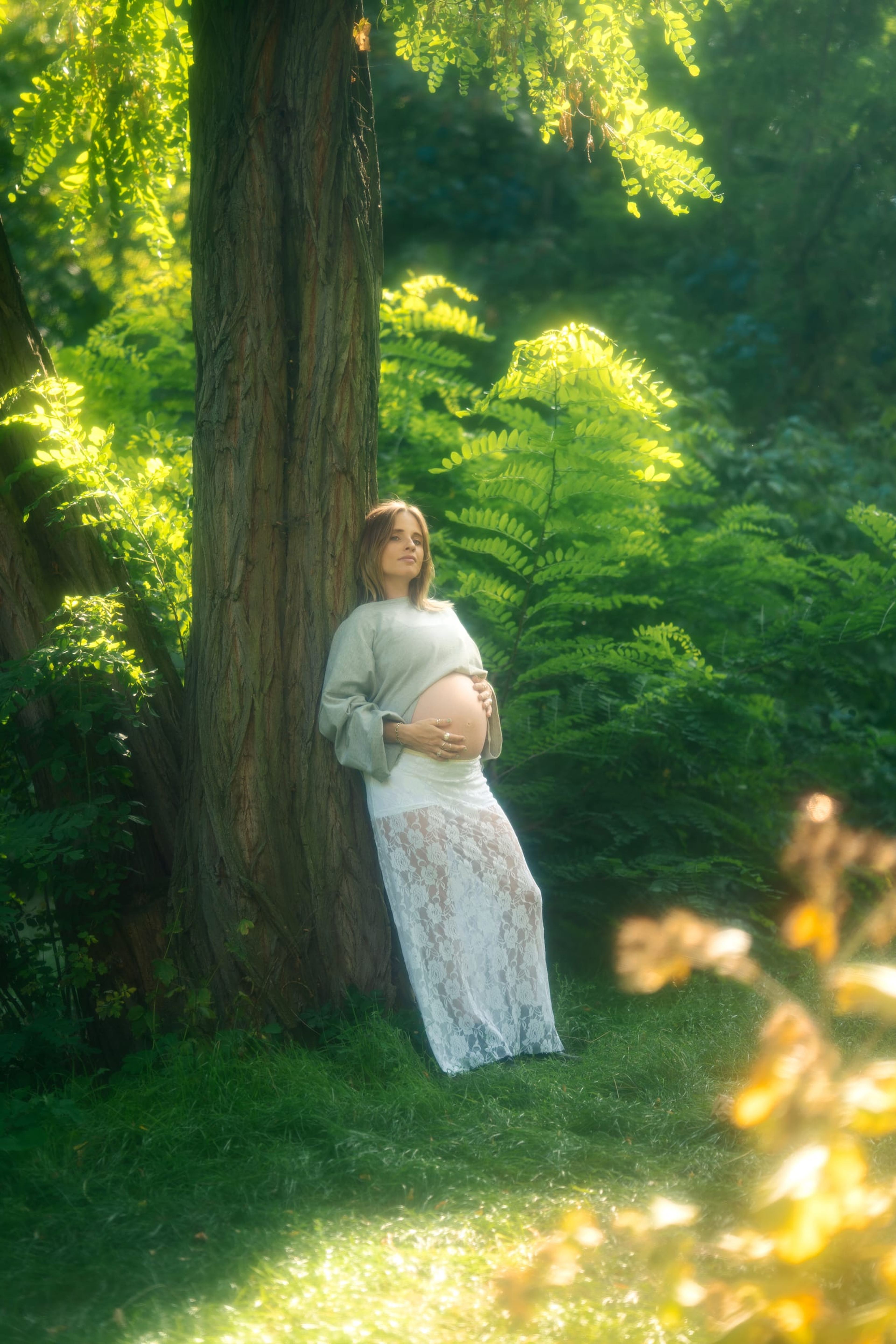 Expectant mother leaning against a large tree, cradling her belly in golden forest light