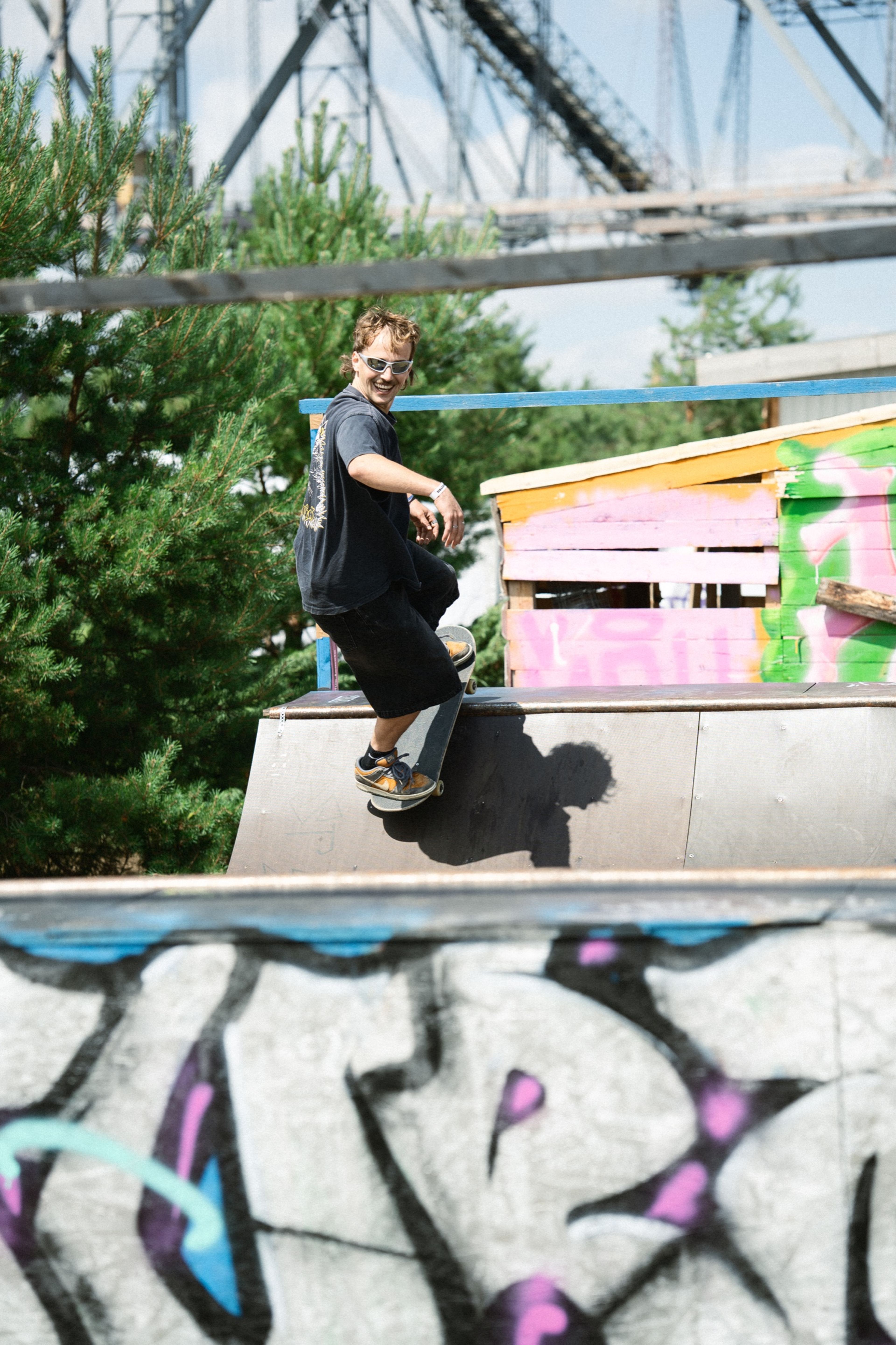 A skateboarder grinning atop a graffiti-covered ramp under a blue sky
