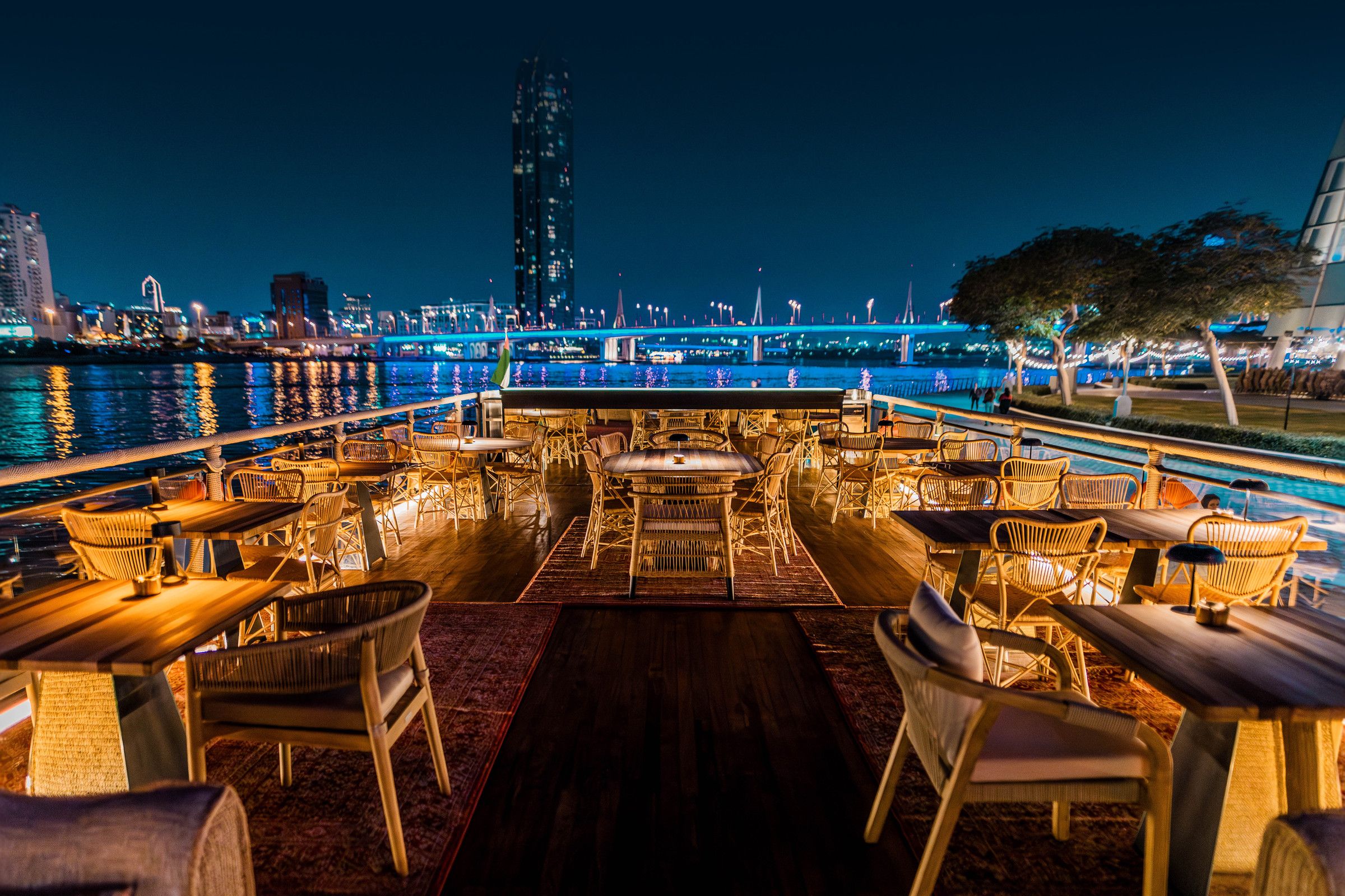 Tables set for dinner on the deck of a dhow, the Dubai skyline lit at night